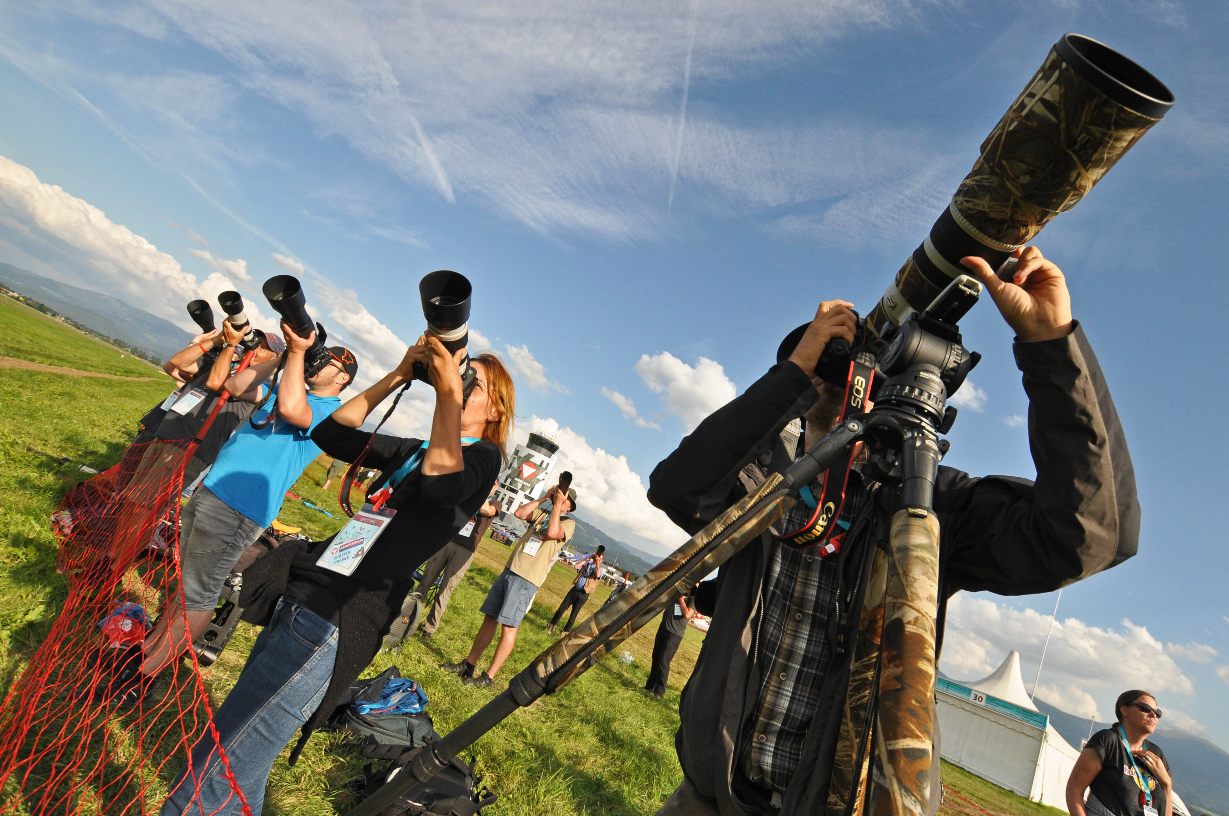 Menschen stehen mit Kameras auf grünem Gras und richten ihre langen Teleobjektive gen Himmel, als würden sie den Himmel fotografieren. Im Vordergrund ist ein rotes Netz zu sehen, während Zelte und Wolken den Hintergrund füllen. Die Gruppe wirkt konzentriert, ihre Kameras stehen auf Stativen, um Stabilität zu gewährleisten.