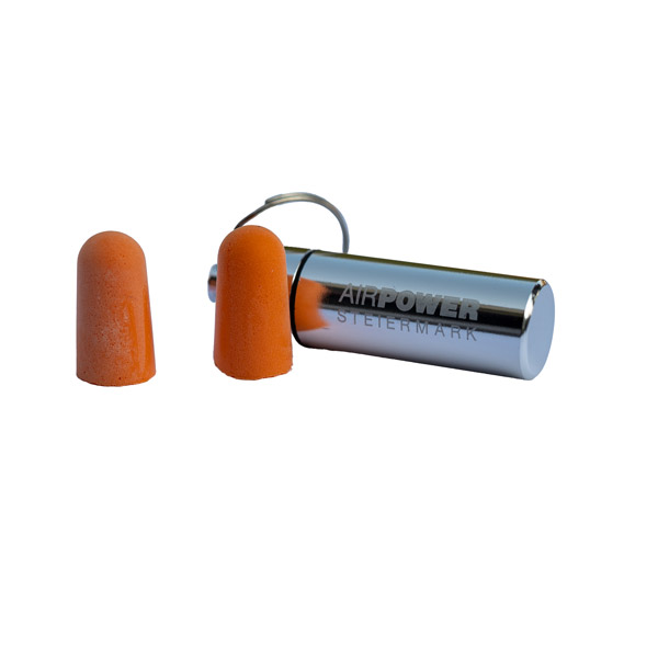 Zwei orangefarbene Ohrstöpsel neben einem silbernen zylindrischen Schlüsselanhängerbehälter mit der Gravur „AIRPOWER“ und „STEIERMARK“.