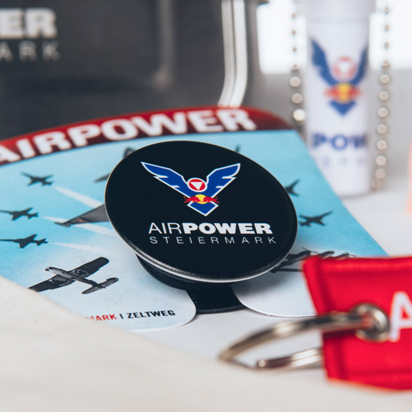 Nahaufnahme von AirPower Steiermark-Artikeln, darunter eine Broschüre, ein Popsocket, ein Schlüsselband, ein Schlüsselanhänger und Lippenbalsam auf einem Tisch.
