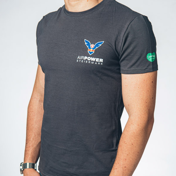 Person in marineblauem „Airpower Steiermark“-T-Shirt mit Logo, von der Schulter bis zur Taille zu sehen, heller Hintergrund.