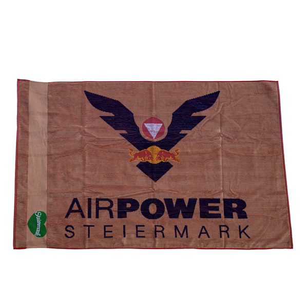 Ein braunes Handtuch mit „AIRPOWER STEIERMARK“ und einem Logo mit einem Adler mit roter Raute und geflügeltem Stier.